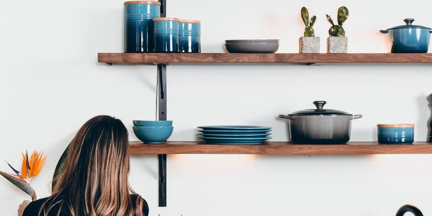 Woman standing beside organized kitchen shelves with cookware and plants.