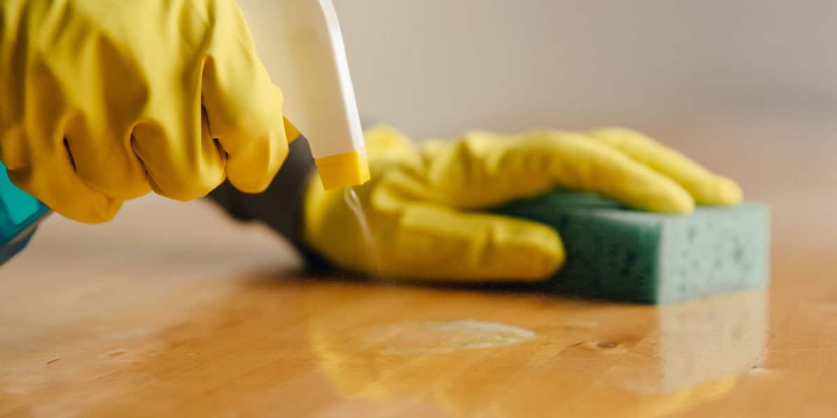 Gloved hands spraying cleaner and wiping a wooden surface with a sponge.
