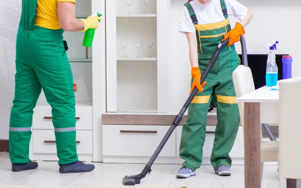 Maid vacuuming living room floor while another cleaner wipes shelves